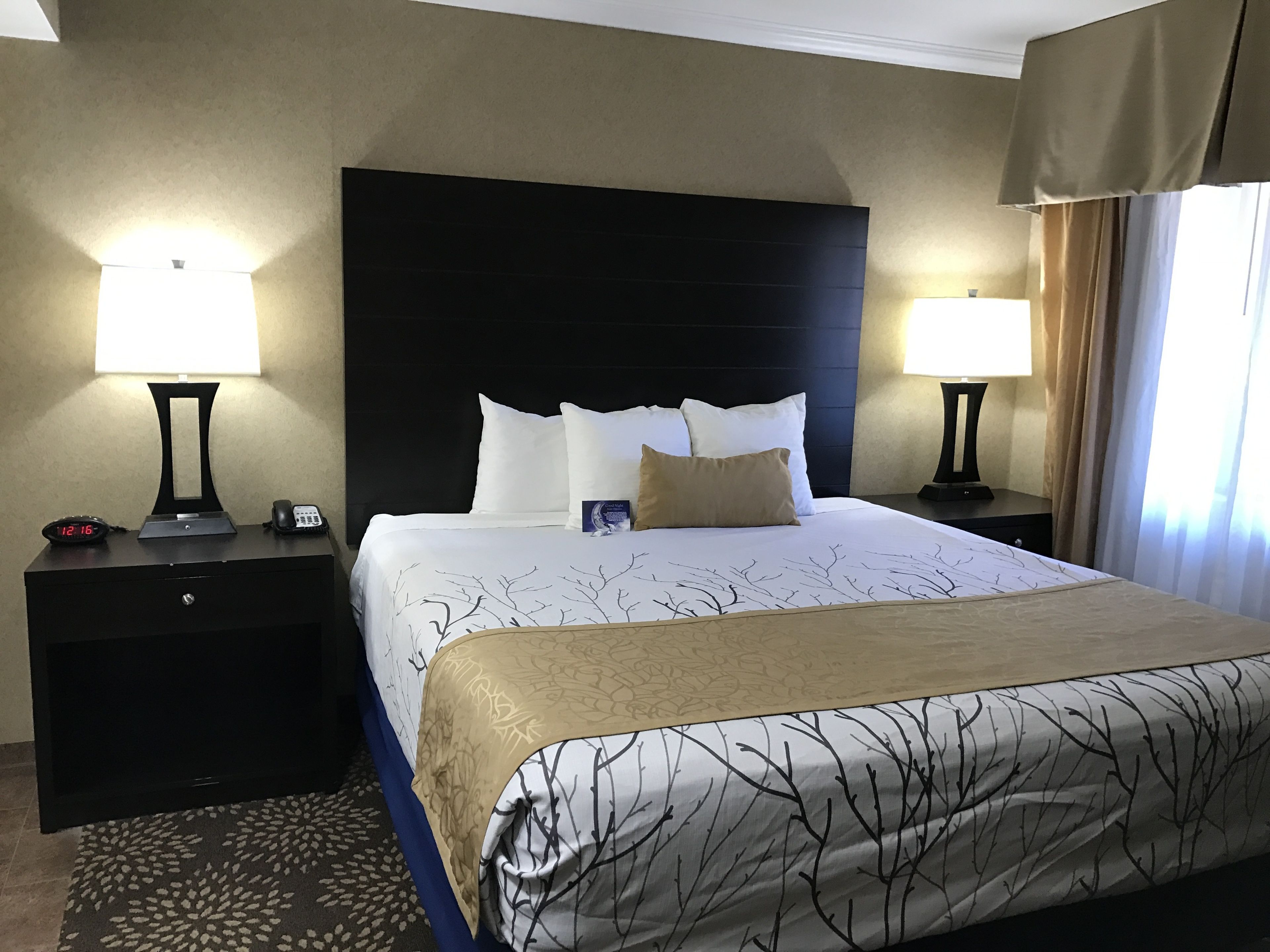 Une chambre d’hôtel avec un grand lit, deux tables de chevet, une commode, une lampe, une horloge et une fenêtre avec des rideaux.