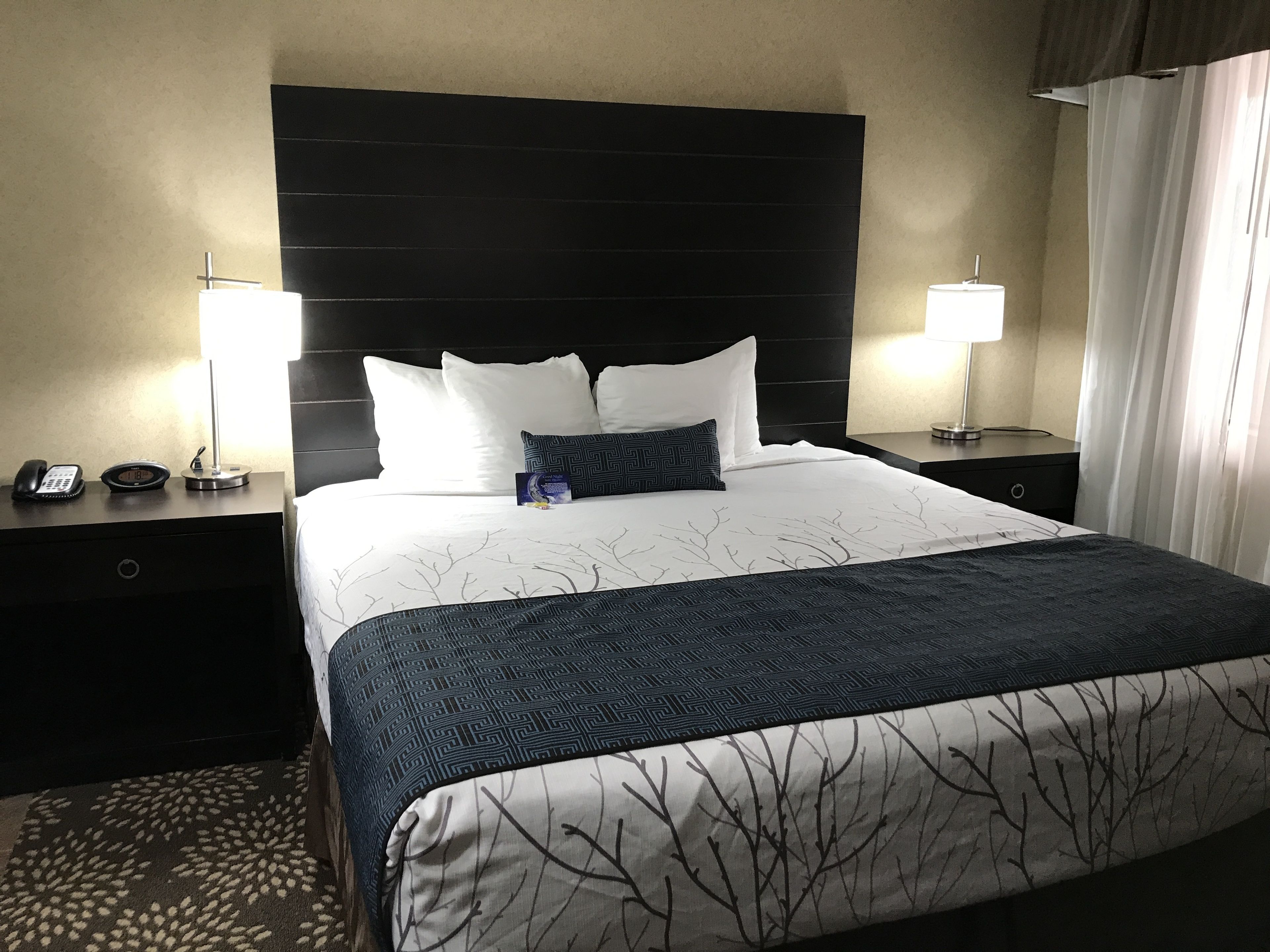 A hotel room with a large bed, two bedside tables, a telephone, a clock, and a lamp on each table.