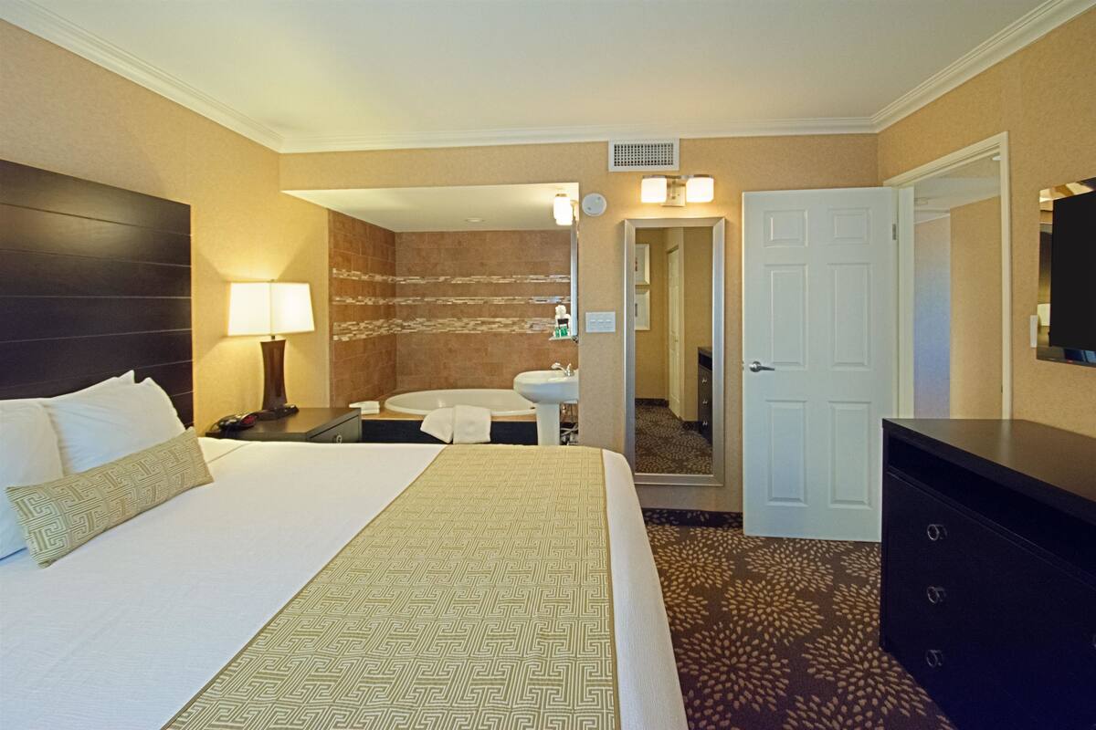 A hotel room with a bed, a nightstand, a lamp, a bathroom with a sink and mirror, and a dresser.