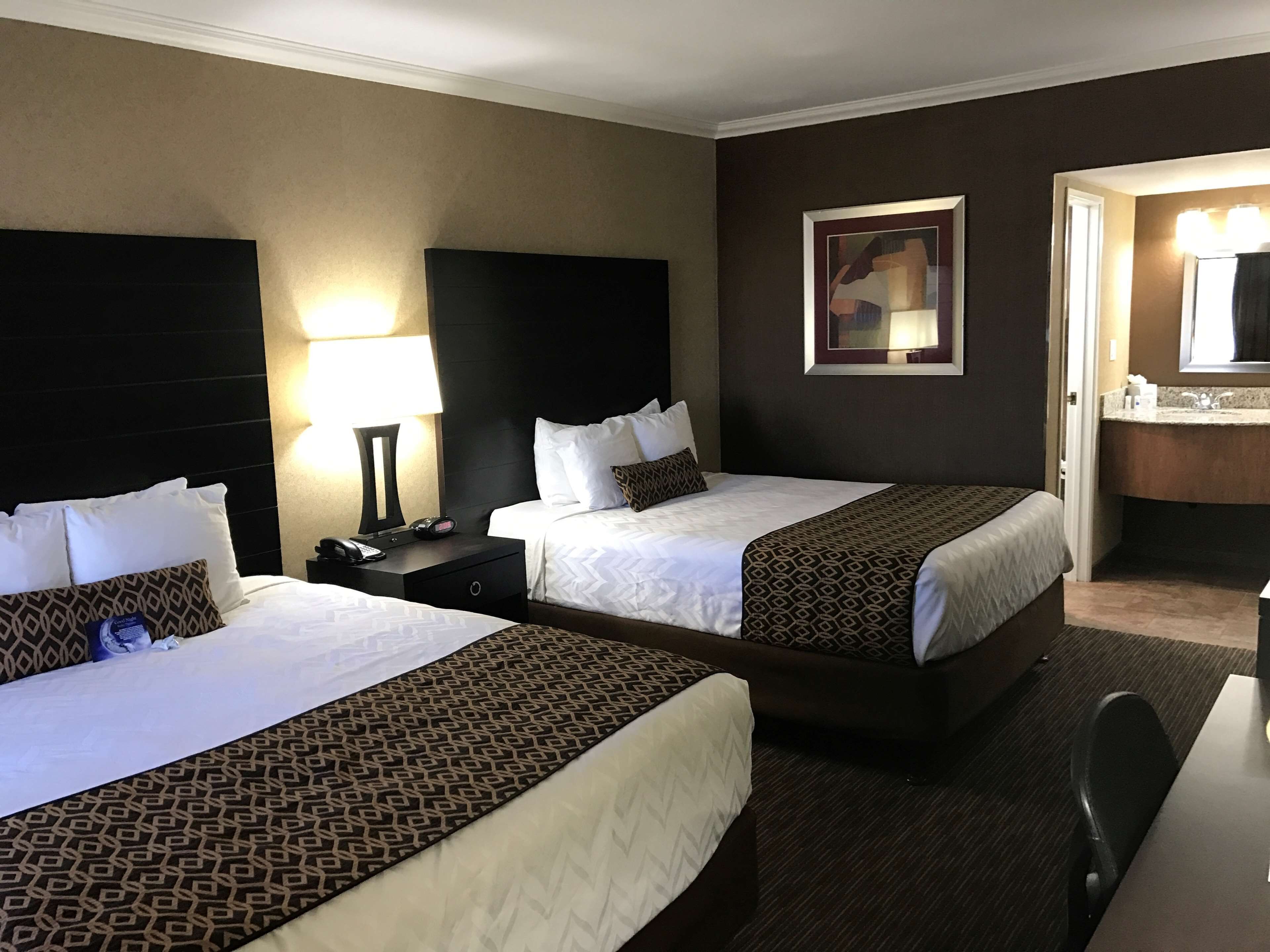 A hotel room with two beds, a nightstand with a lamp, a bedside table, a mirror, and a bathroom with a sink and mirror.