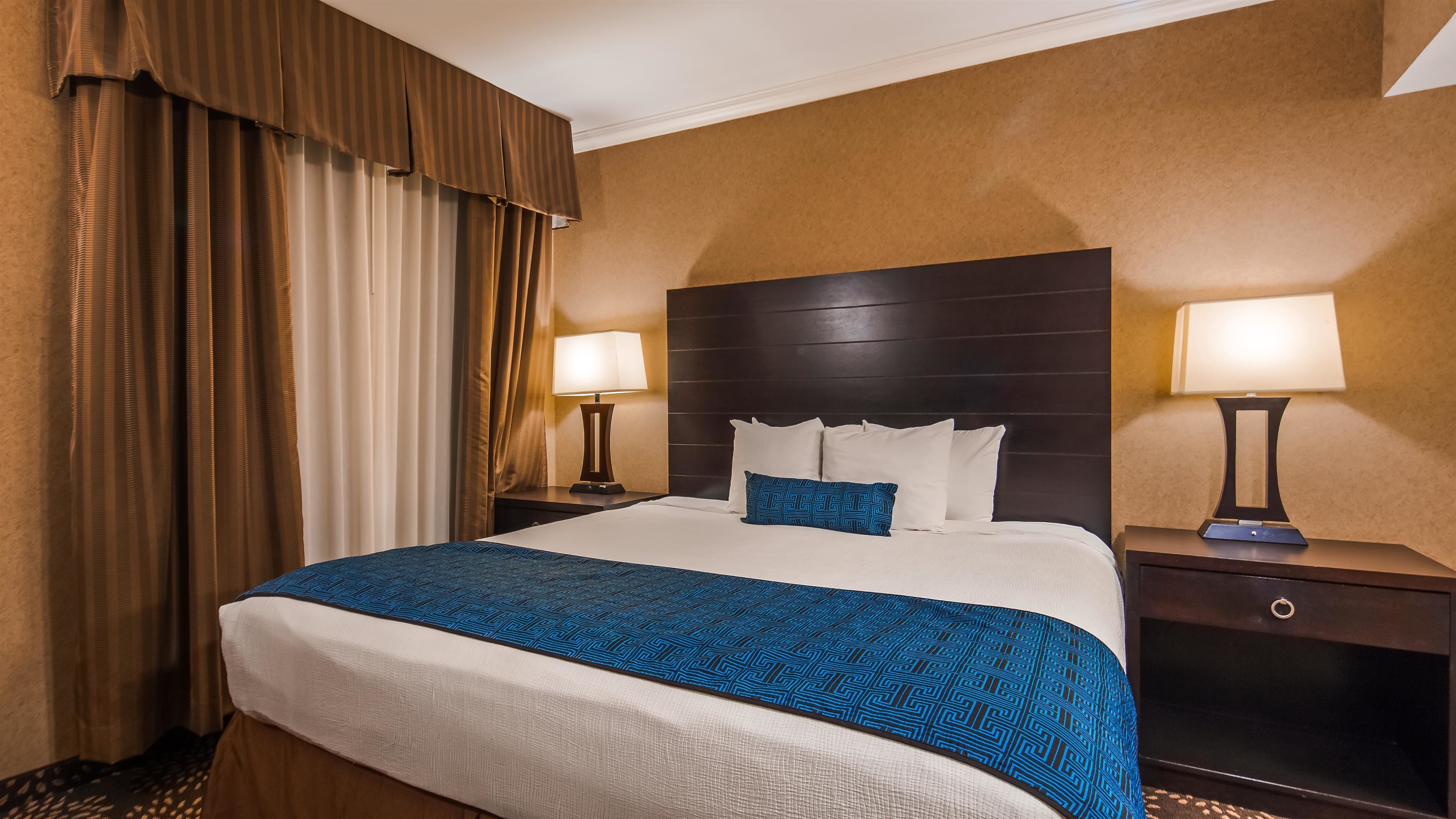 A hotel room with a large bed, two bedside tables, a headboard, and curtains.