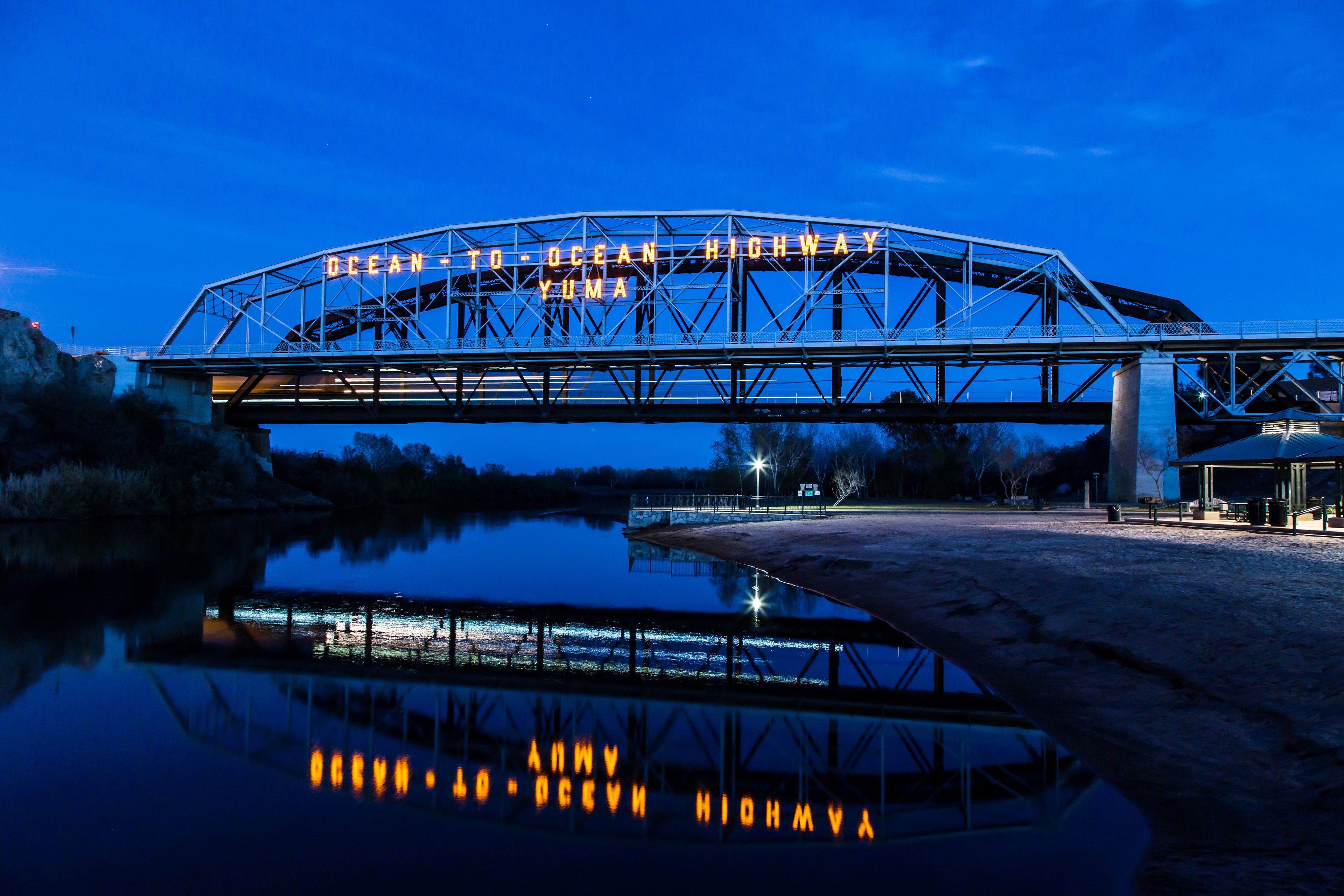 A metal bridge with the inscription "OCEAN TO OCEAN HIGHWAY YUMA" spanning over a body of water, with a reflection visible in the water.