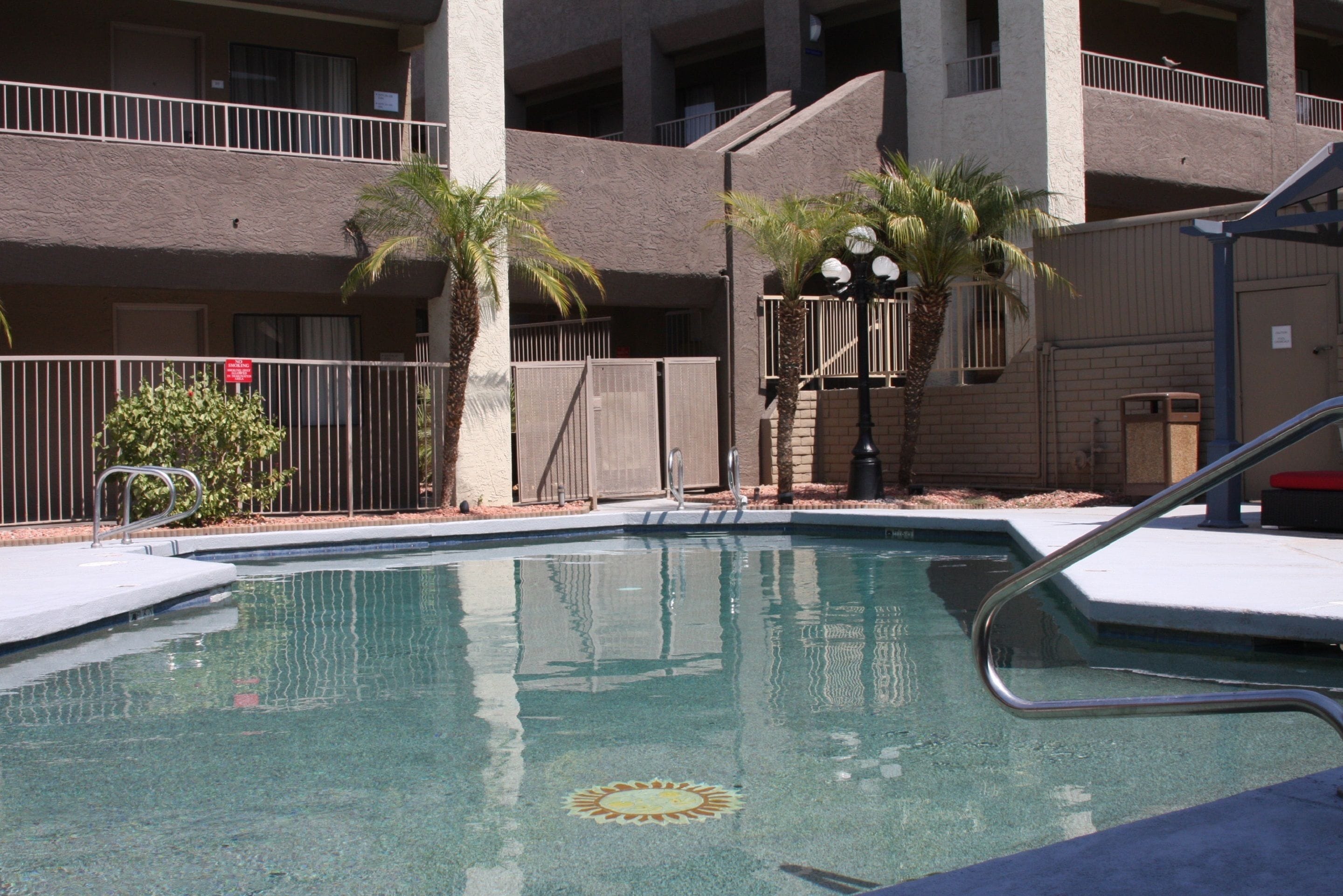 A swimming pool with a sun design, surrounded by palm trees and a multi-story building with balconies.