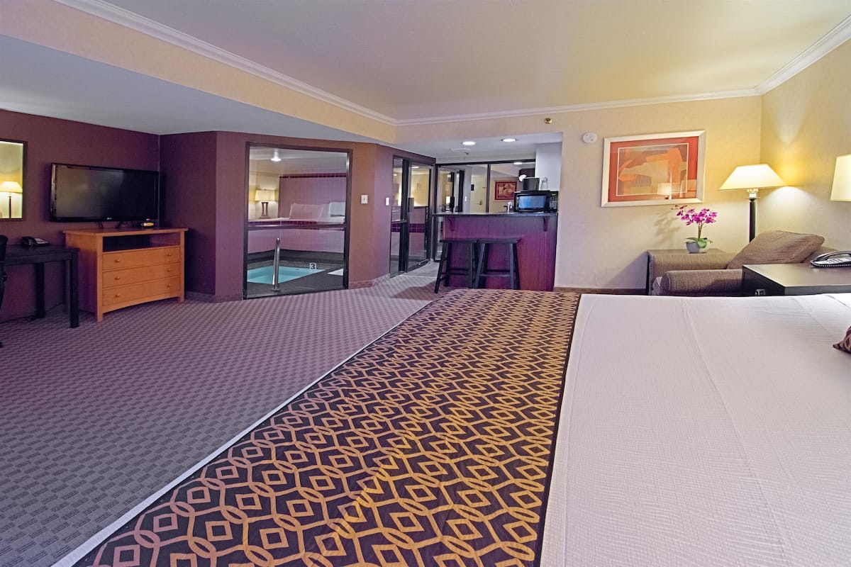 A hotel room with a bed, a TV, a desk with a chair, a small bar area, and a bathroom visible through a glass door.