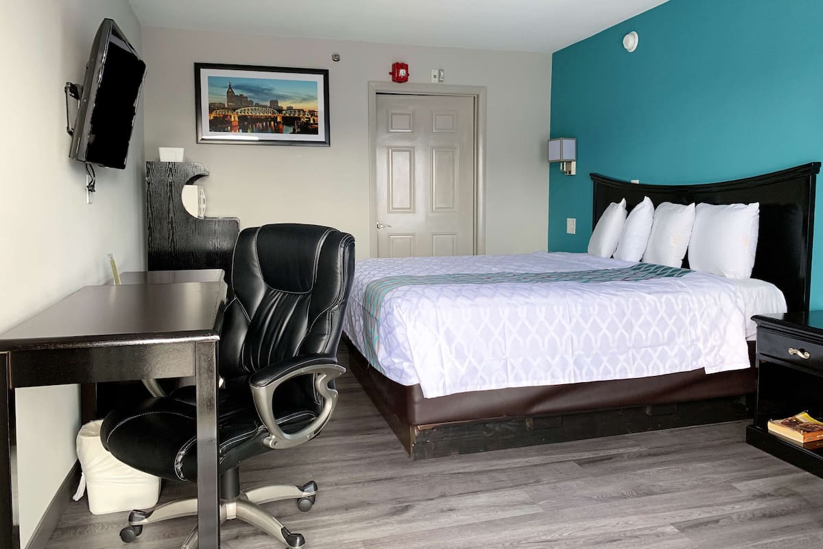 A hotel room with a bed, a desk, a chair, a TV, and a painting on the wall.