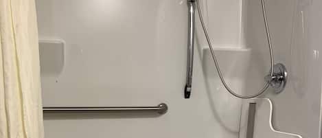 A bathroom with a white shower enclosure, grab bars, a handheld showerhead, and a floor drain.