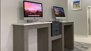 Dos computadoras Apple iMac sobre un escritorio blanco con un cajón, frente a una pared gris con un cuadro enmarcado.
