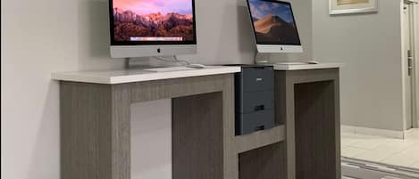 Two Apple iMac computers on a white desk with a drawer, set against a grey wall with a framed picture.
