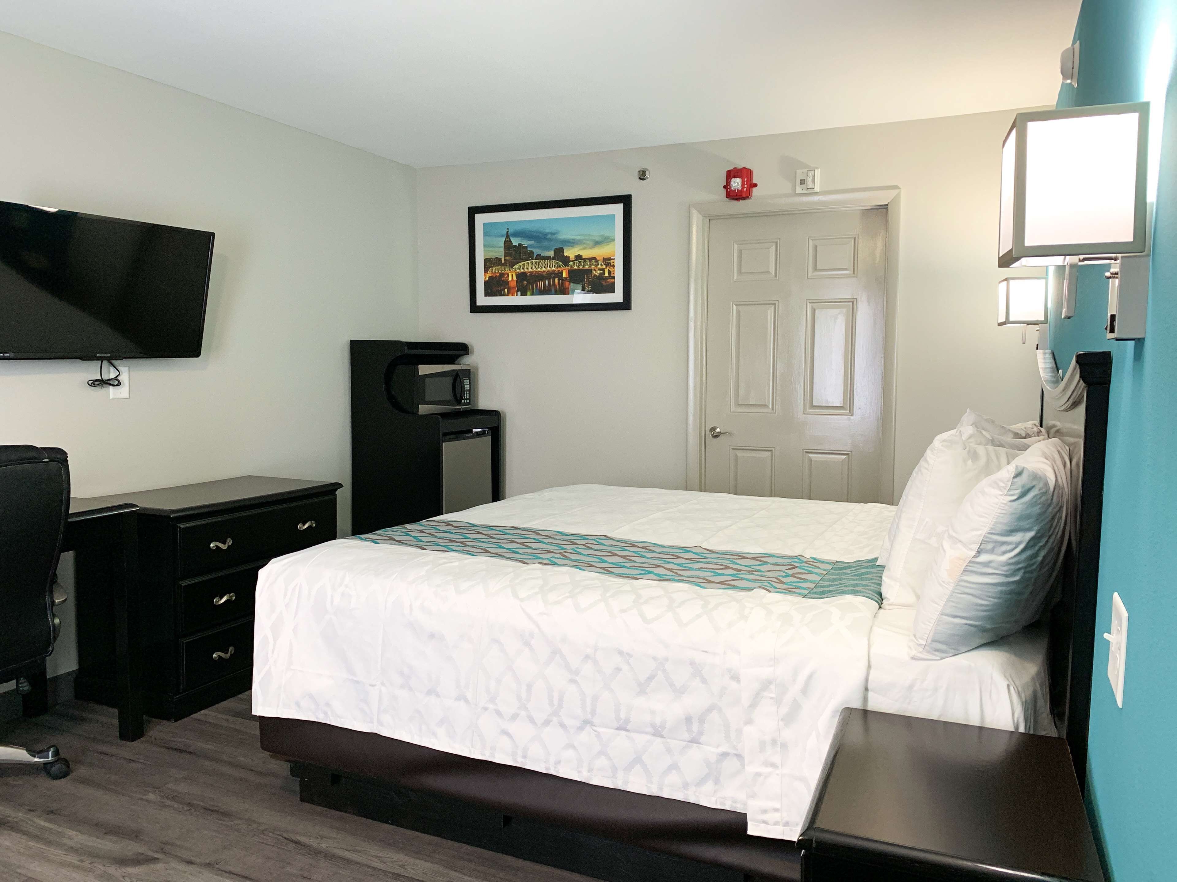 A hotel room with a bed, a desk with a chair, a TV mounted on the wall, a microwave, and a door with a red emergency light.
