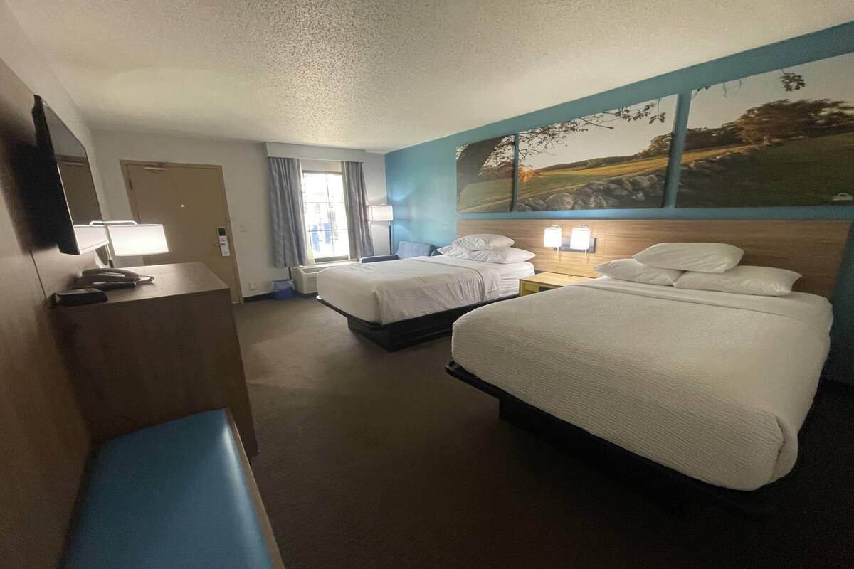 A hotel room with two beds, a desk with a lamp, a bench, and a large window with curtains.