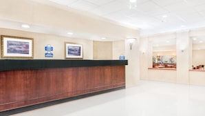 A reception area with a wooden reception desk, framed pictures on the wall, and a glimpse of a kitchen area in the background. - Days Inn & Suites by Wyndham York (York)