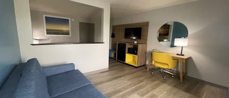 A living room with a blue sofa, a yellow chair, a wooden desk, a TV, and a mirror on the wall.