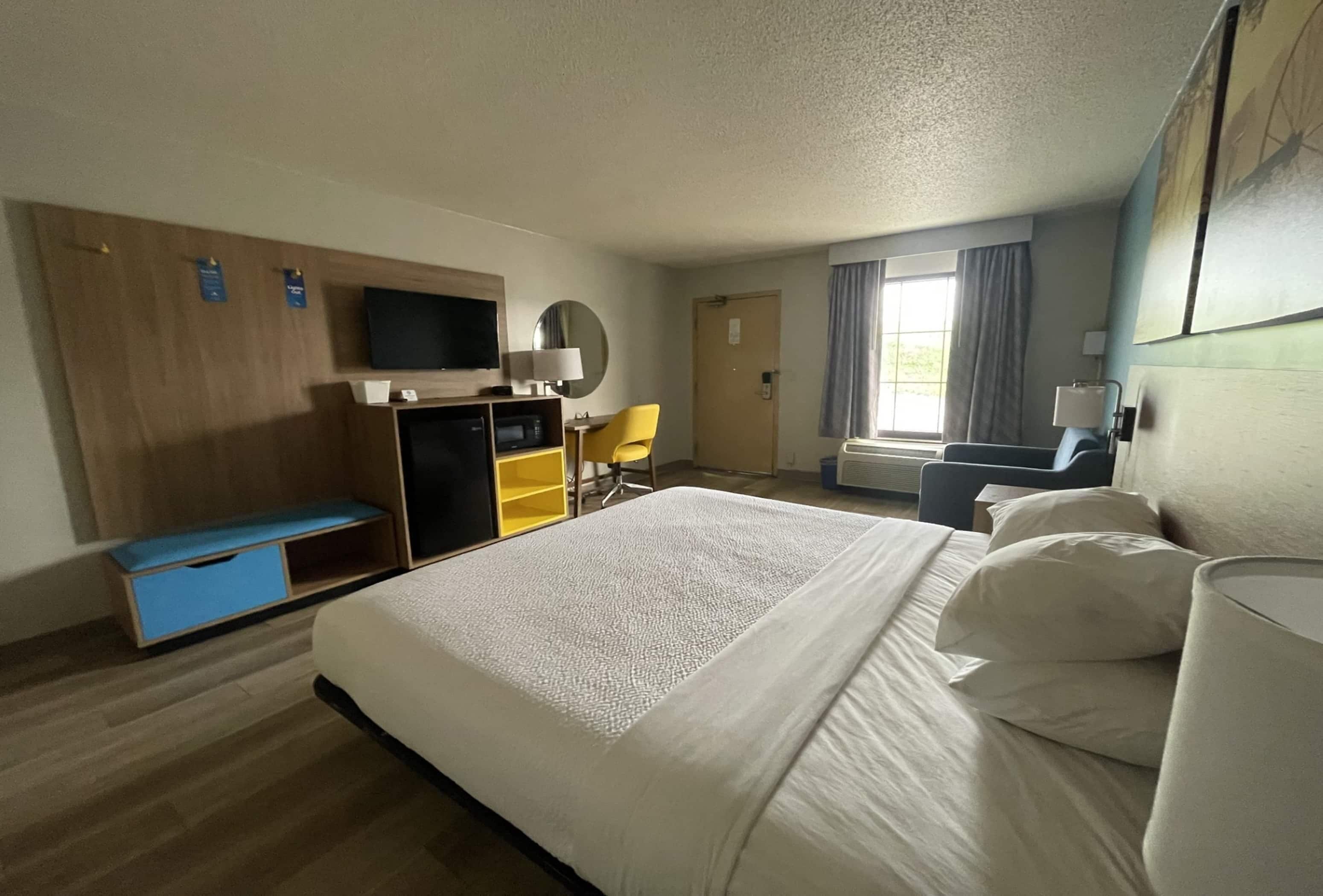 Habitación de hotel con una cama grande, un escritorio con computadora, una silla, un televisor y una ventana con cortinas.
