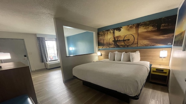 A hotel room with a large bed, a desk, a mirror, and a wall with a painting. - Days Inn & Suites by Wyndham York (York)