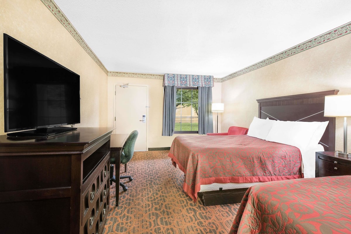A hotel room with a TV, a desk, two beds, a chair, and a window with curtains.