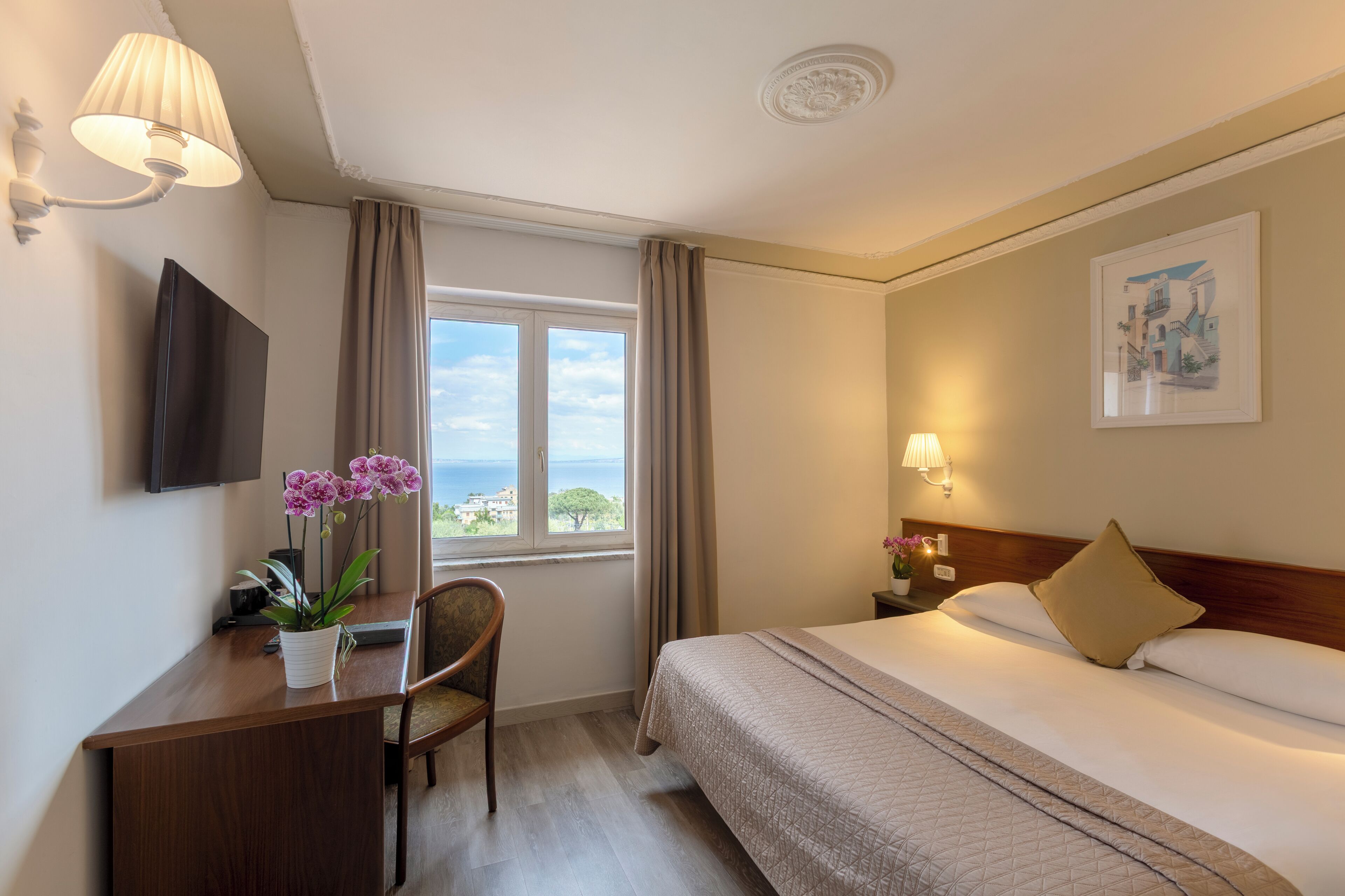 A hotel room with a bed, a desk with a chair, a television, a window with curtains, and a wall-mounted lamp.