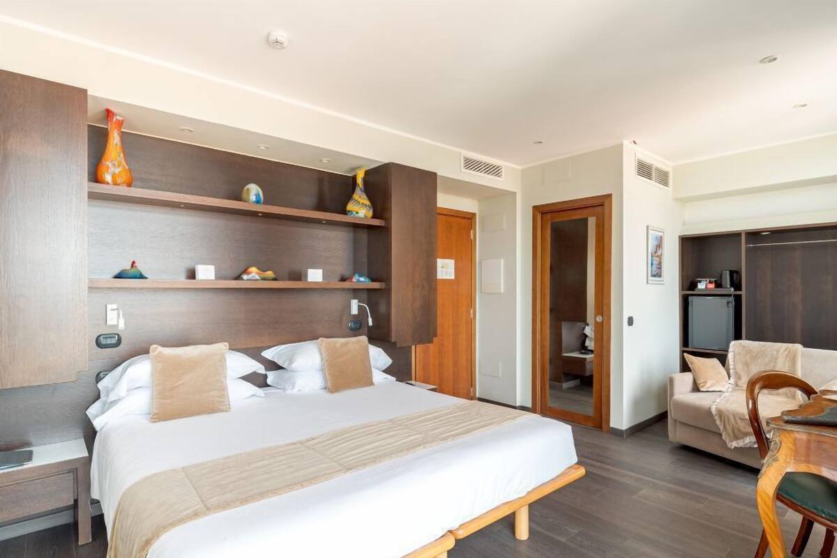 A hotel room with a large bed, wooden headboard, bedside tables, a desk with a chair, and a wardrobe.