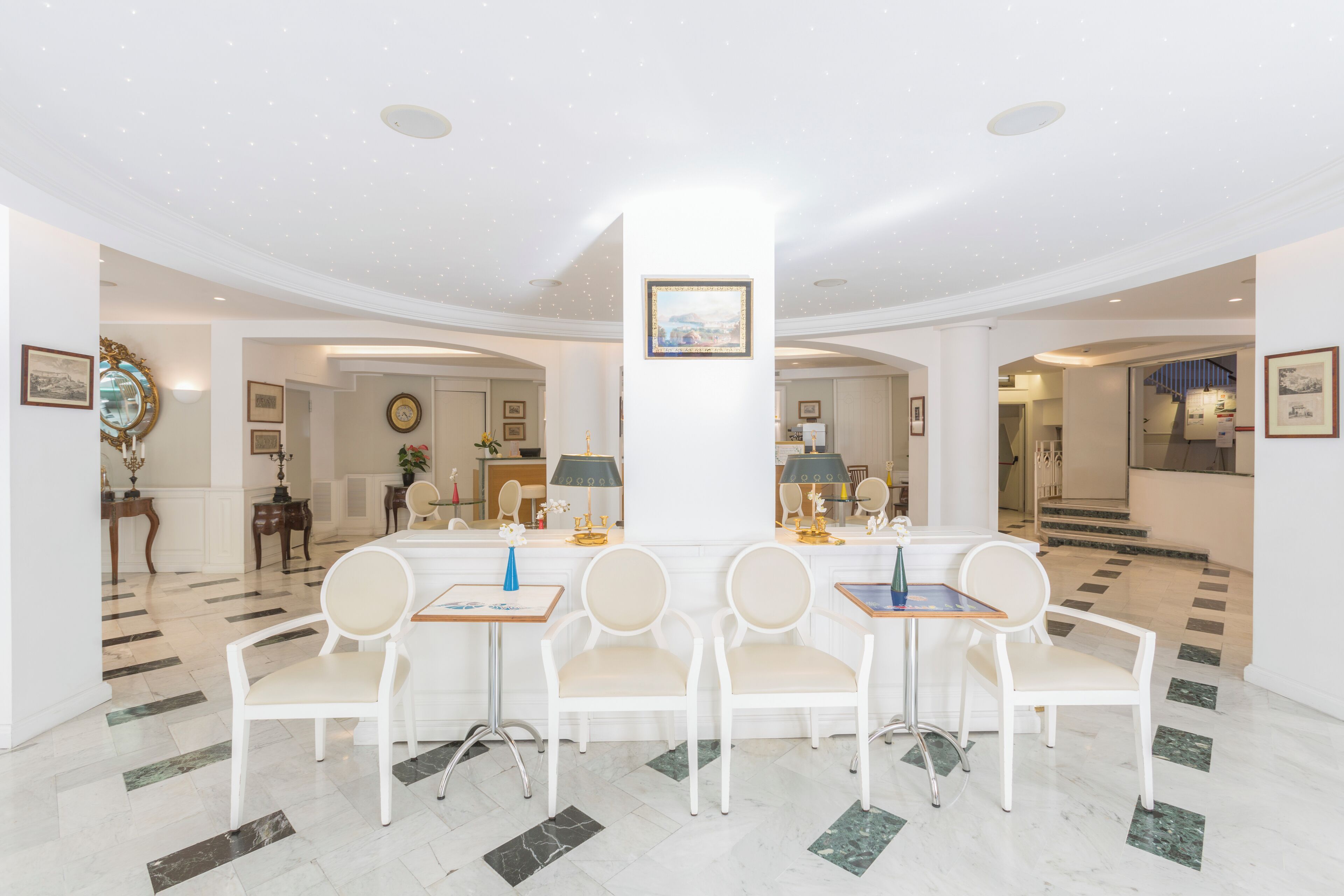 A spacious lobby with a marble floor, white chairs, a central table, and framed artwork on the walls.