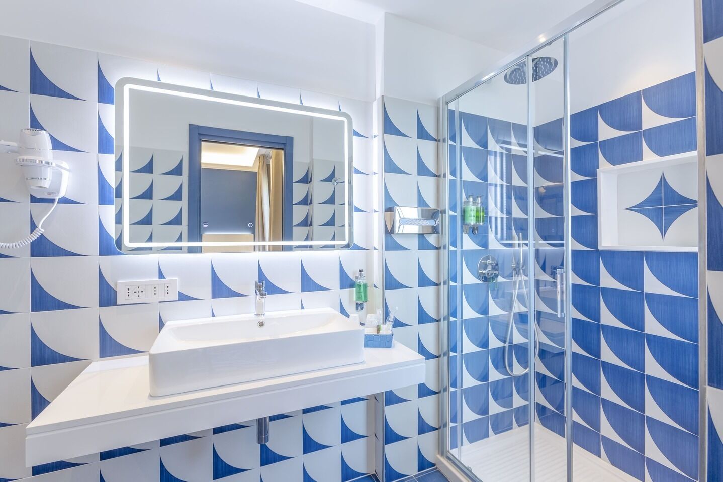 Ein modernes Badezimmer mit geometrischem Fliesenmuster, einem weißen Waschbecken, einer Glasdusche und einem Spiegel mit integrierter Beleuchtung.