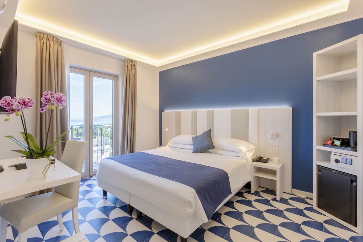 A hotel room with a bed, bedside table, nightstand, and a small kitchenette.
