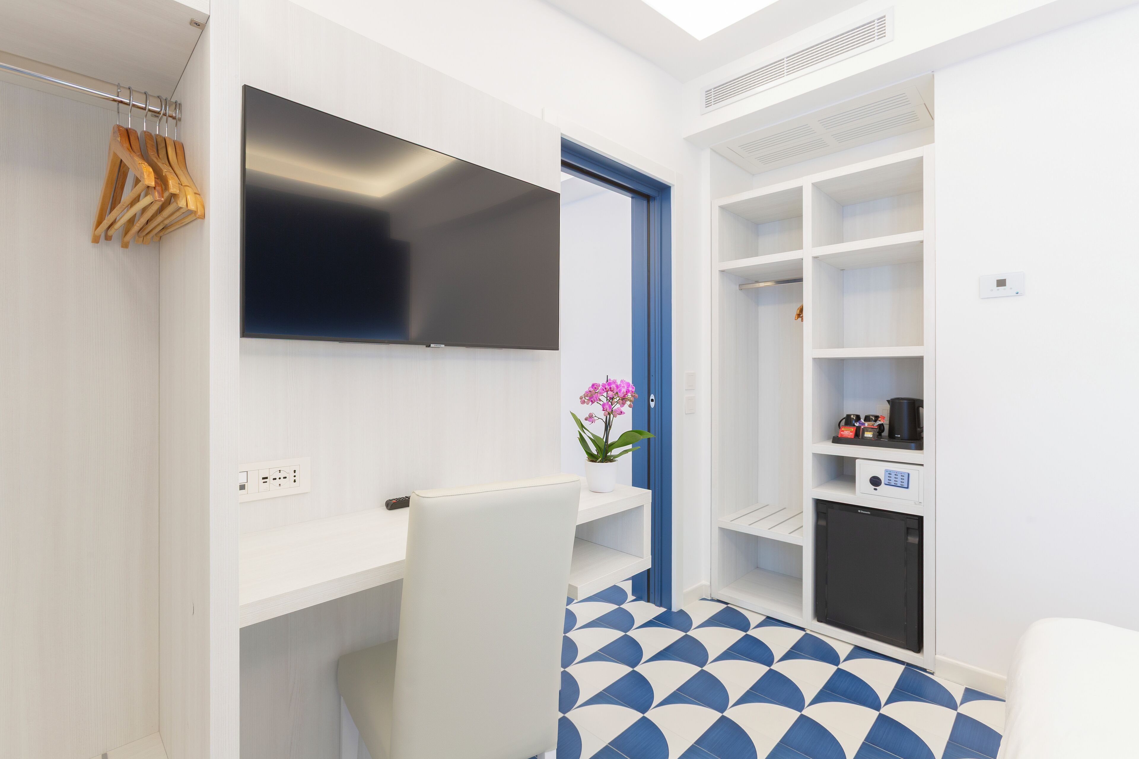 A modern room with a flat-screen TV, a desk, a chair, a wardrobe, and a small kitchenette.