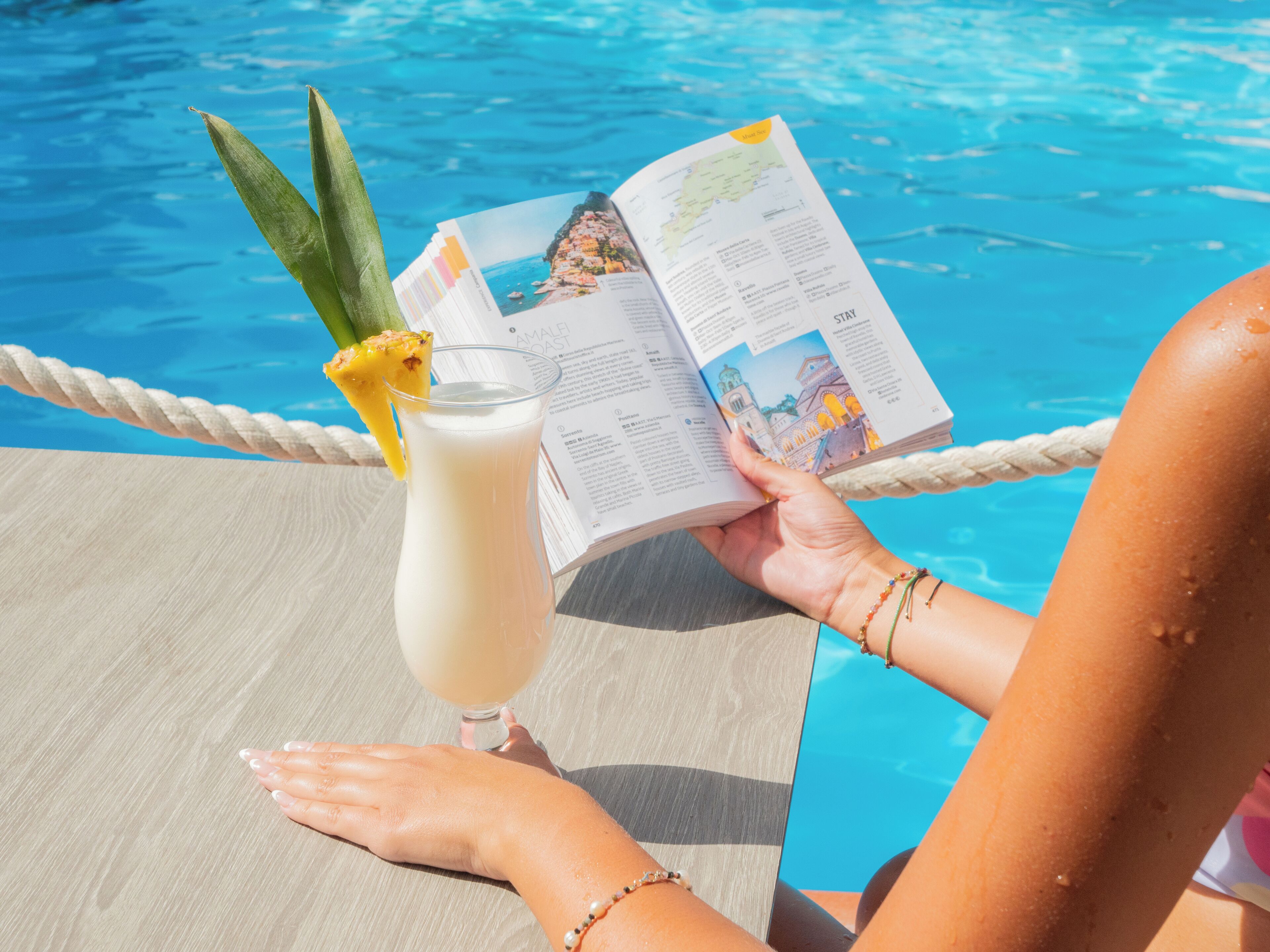A person reading a magazine by a poolside with a drink.
