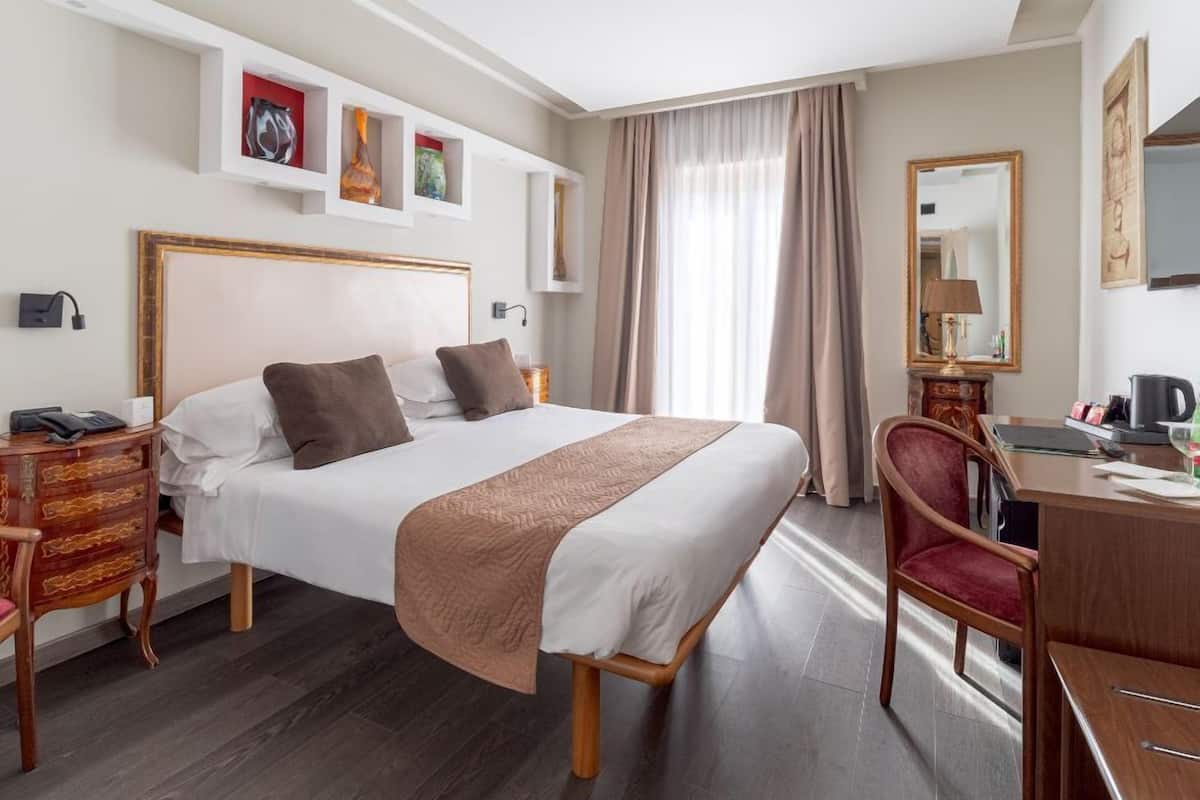 A hotel room with a bed, bedside table, chair, desk, and a mirror.