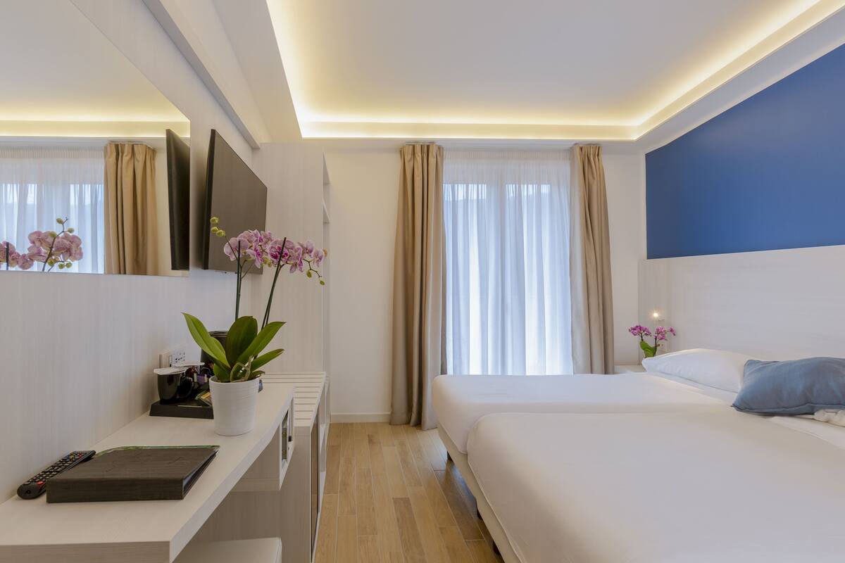 A modern hotel room with a large bed, a desk with a computer and a plant, a flat-screen TV, and a large window with curtains.