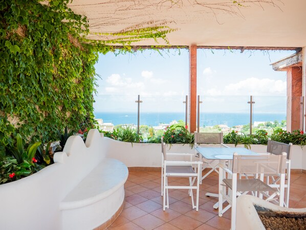 Una terraza con un banco blanco curvo, una mesa y sillas, con vistas a la costa.