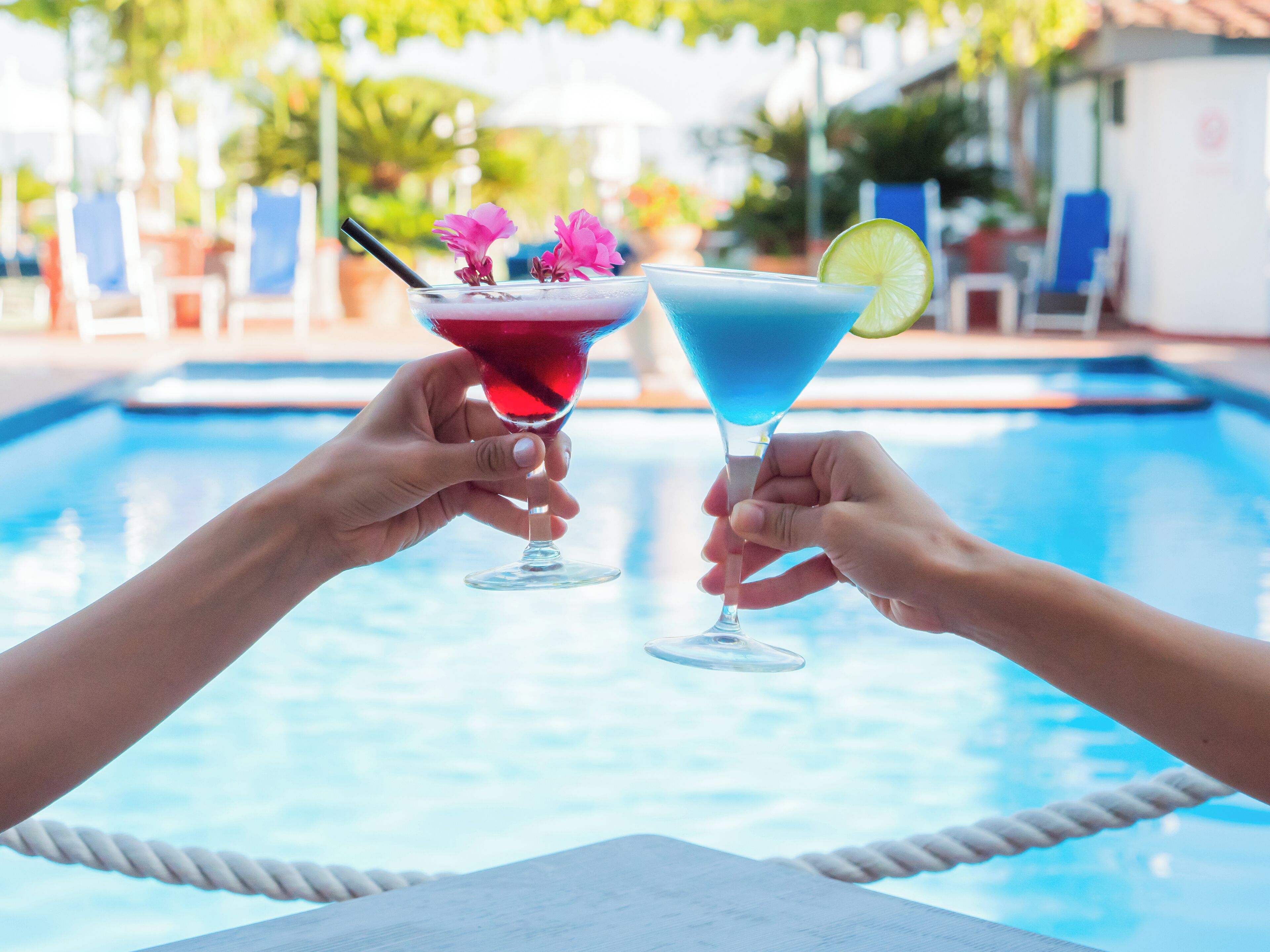 Dos manos sosteniendo copas de martini con bebidas y adornos coloridos junto a una piscina.