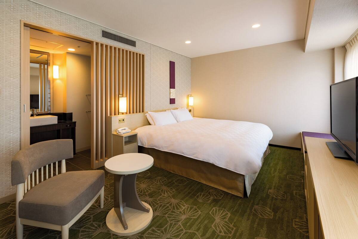 A hotel room with a bed, a desk, a chair, a television, and a bathroom with a mirror and sink.