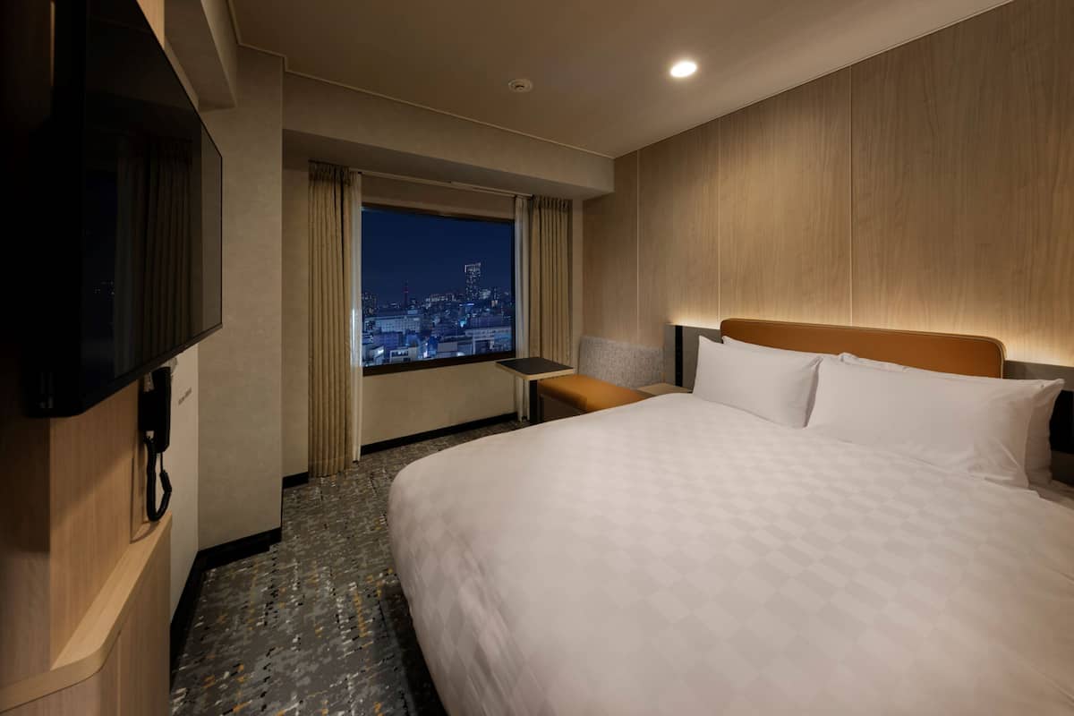 A hotel room with a large bed, a TV, a desk, and a window with a city view.