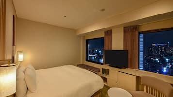 Hotelkamer met een groot bed, een televisie, een bureau met stoel en uitzicht op een stadsbeeld 's nachts.