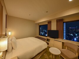 Hotellihuone, jossa on suuri sänky, televisio, työpöytä tuolilla ja näkymä kaupungin yömaisemaan.