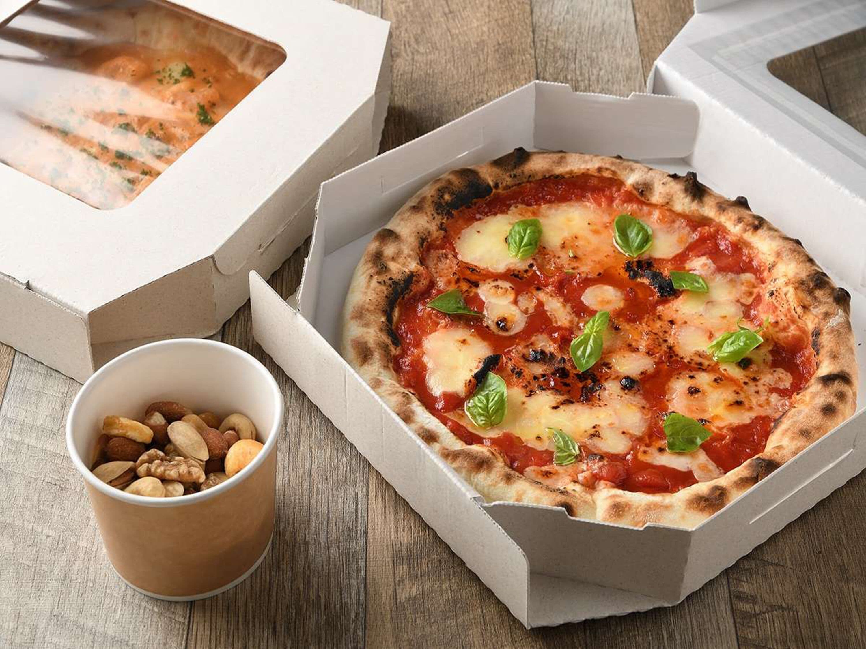 A pizza in a box with a small bowl of nuts beside it.