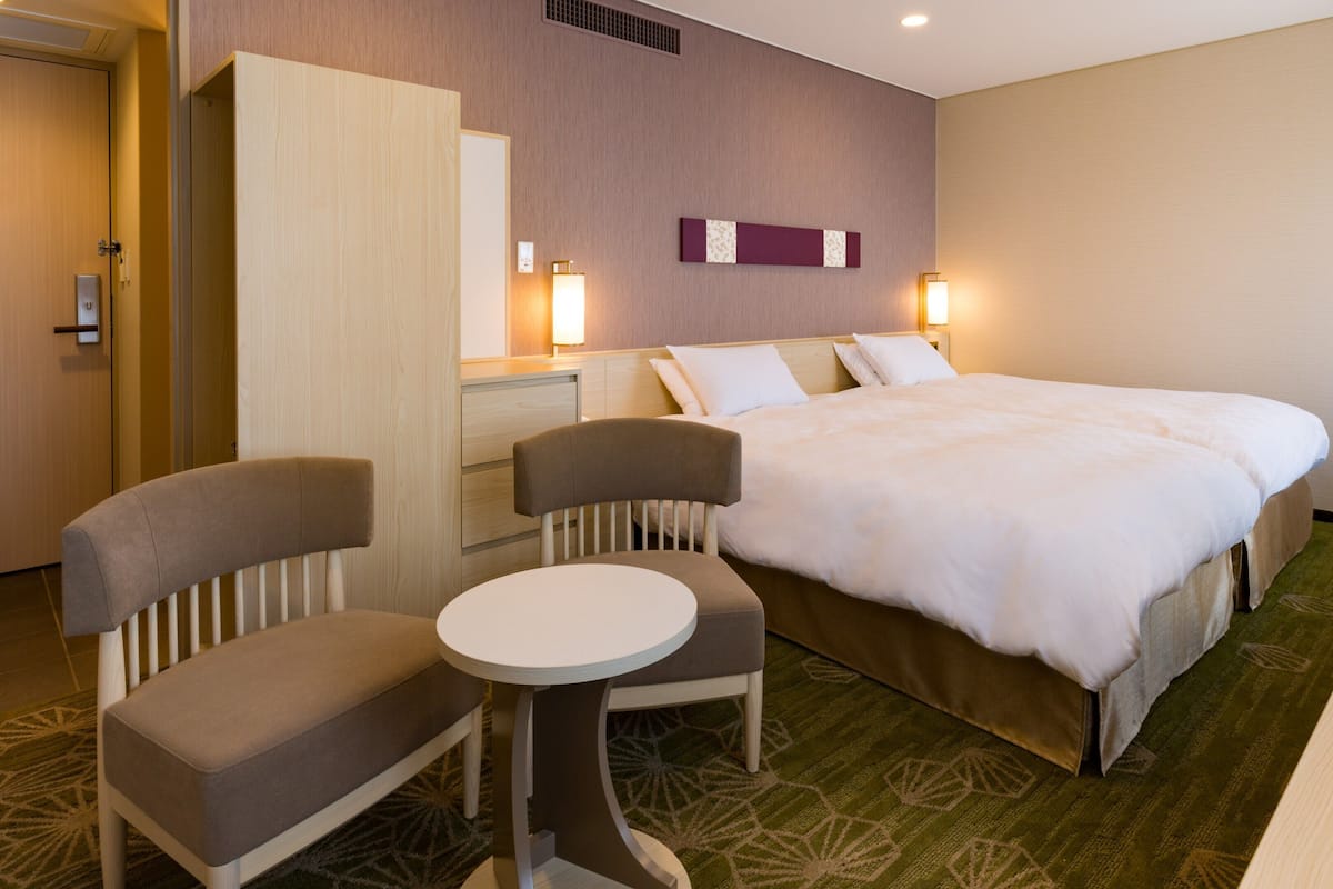 A hotel room with a bed, two chairs, a small table, a dresser, and a mirror.