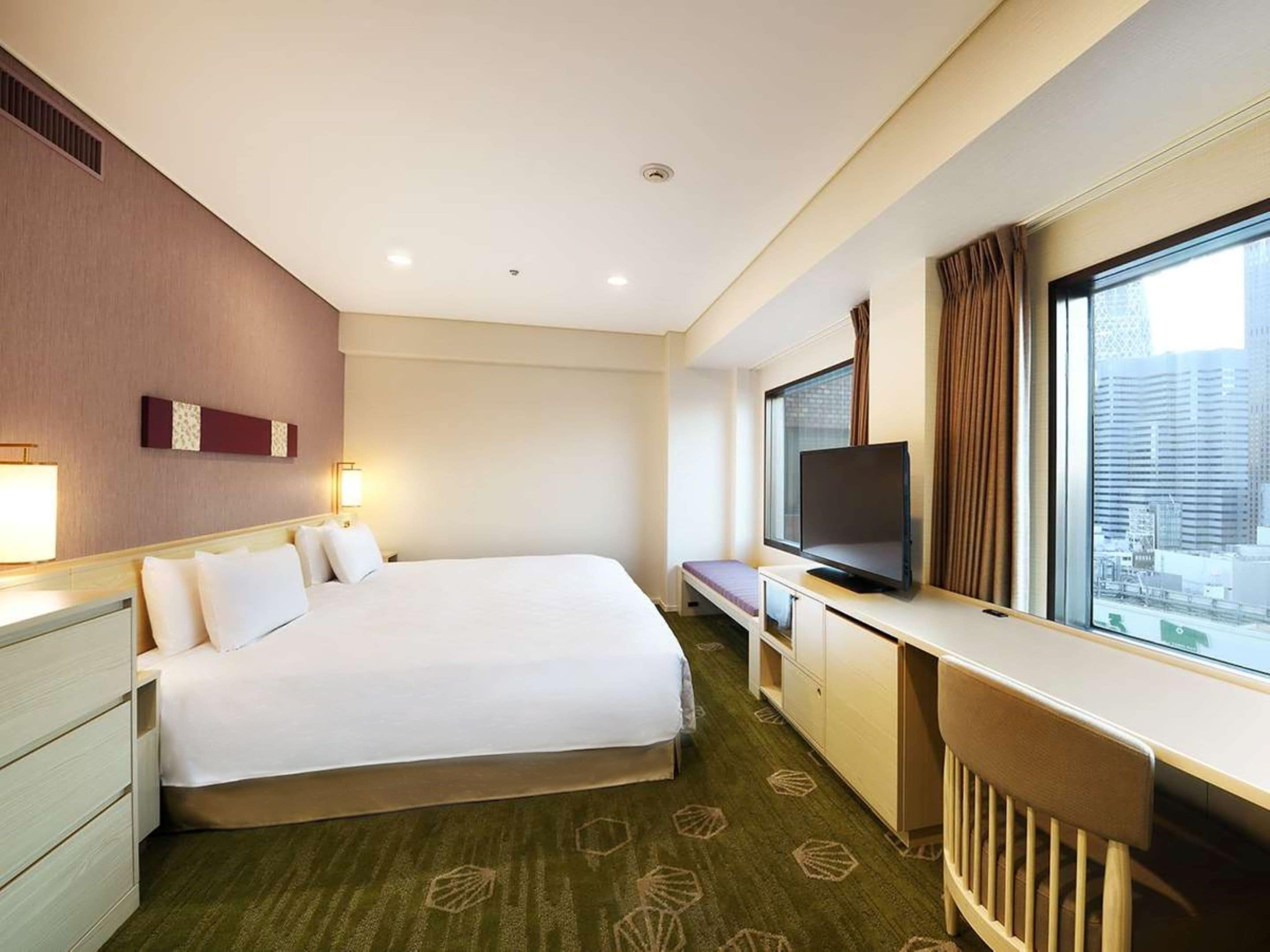 A hotel room with a large bed, a desk with a TV, a window with curtains, and a city view.