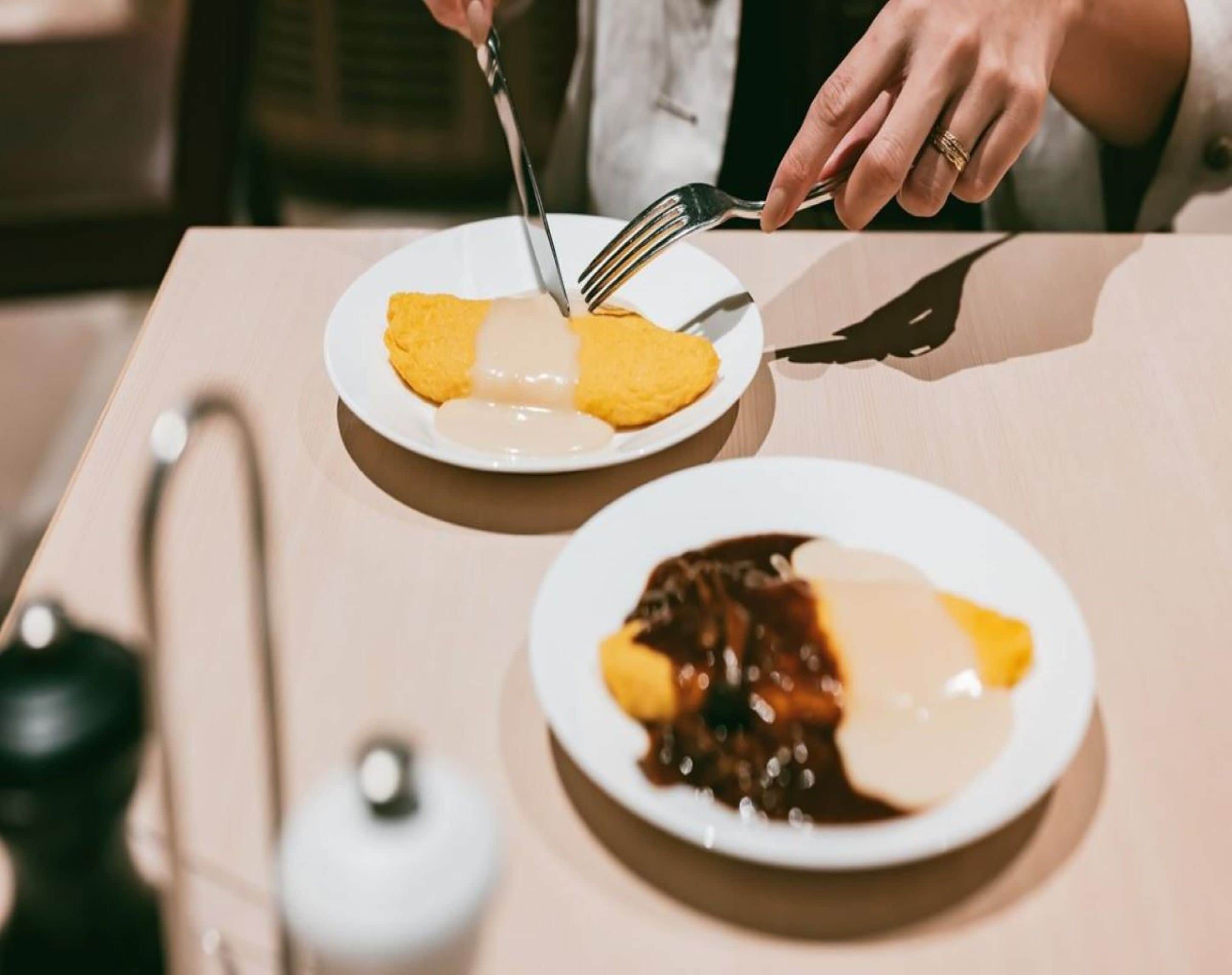 A dining table with two plates of food, one with a yellow dessert and the other with a chocolate dessert, and a person using a fork.