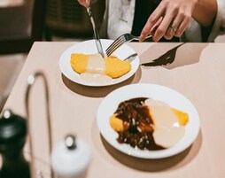 Ruokapöydällä on kaksi lautasellista ruokaa: toisella on keltainen jälkiruoka ja toisella suklaajälkiruoka, ja pöydässä istuva henkilö käyttää haarukkaa.