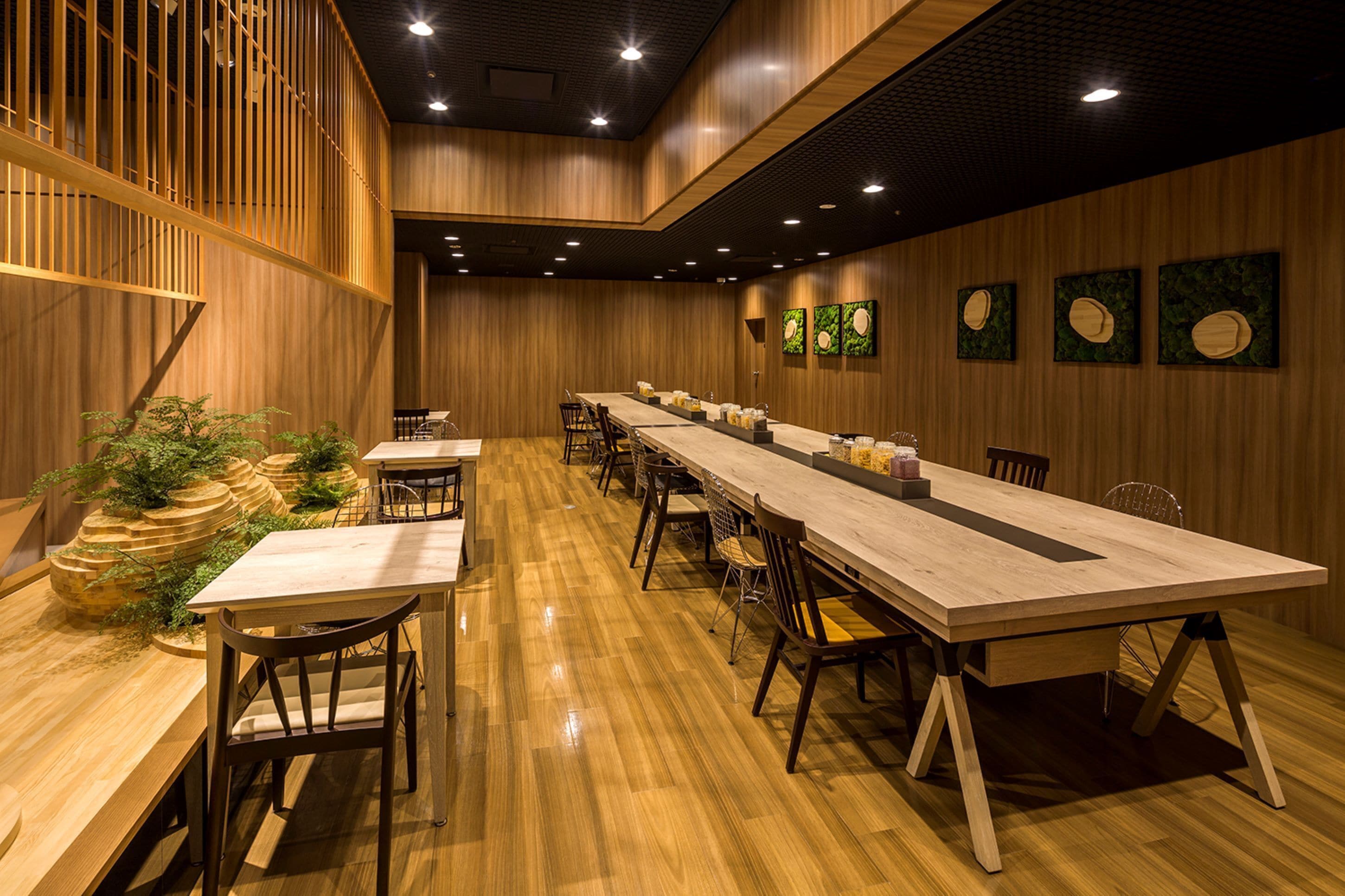Une longue table à manger en bois, entourée de chaises, avec des murs et un plafond recouverts de panneaux en bois, et une plante en pot placée sur le côté gauche.