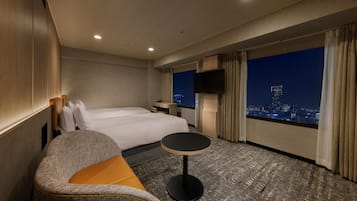 Ein Hotelzimmer mit einem Bett, einer Couch, einem kleinen Tisch und Blick auf die Stadt durch große Fenster.