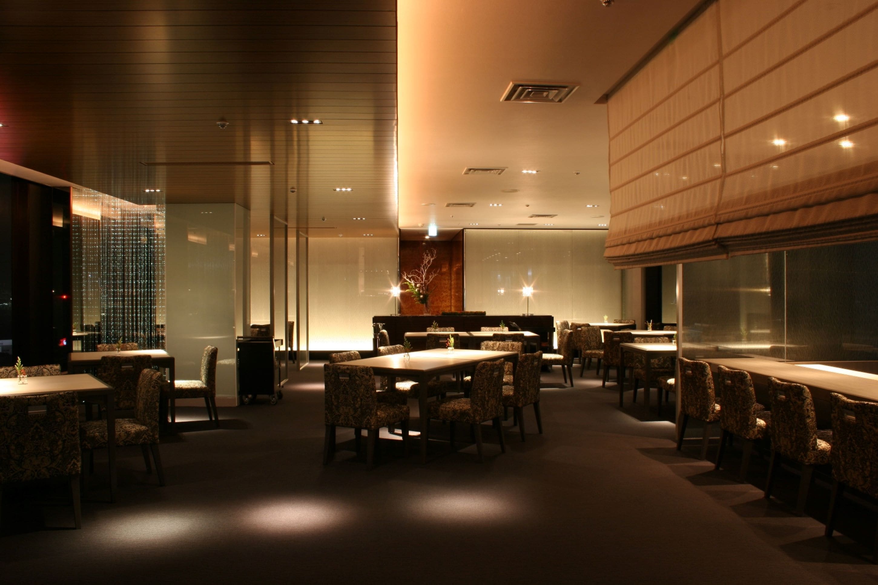 A modern dining area with multiple tables and chairs, a large window with a view, and a decorative plant in the center.