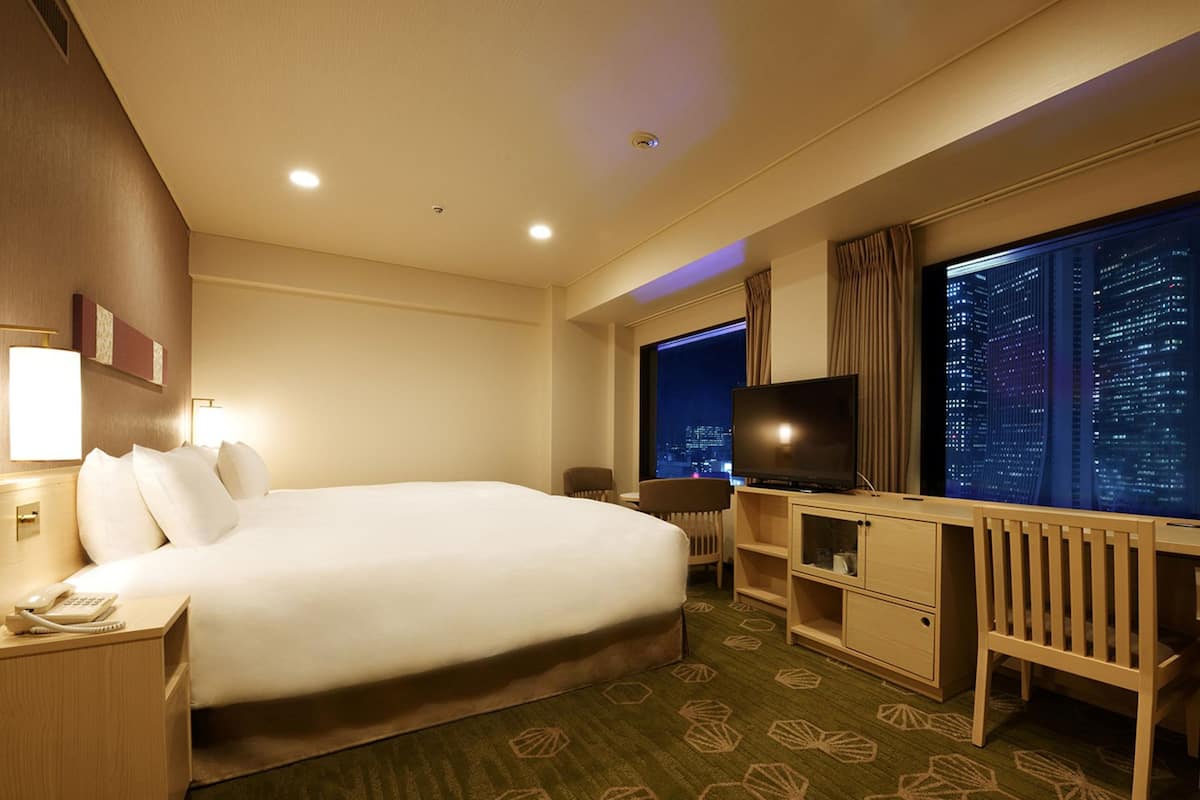 A hotel room with a large bed, a desk with a television, a city view, and a carpet with a geometric pattern.