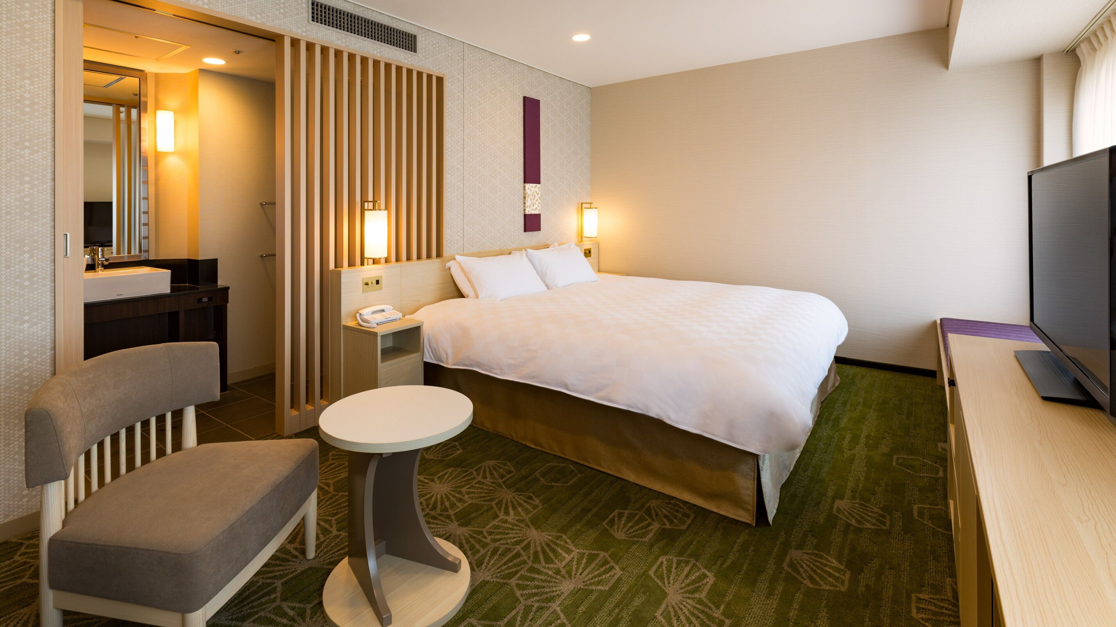 A hotel room with a bed, a desk, a chair, a small table, a TV, and a bathroom with a mirror and sink.