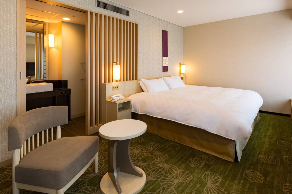 A hotel room with a bed, a desk, a chair, a small table, a TV, and a bathroom with a mirror and sink.