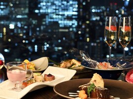 A dining table with various dishes, a glass of pink drink, and two glasses of champagne, set against a cityscape background.