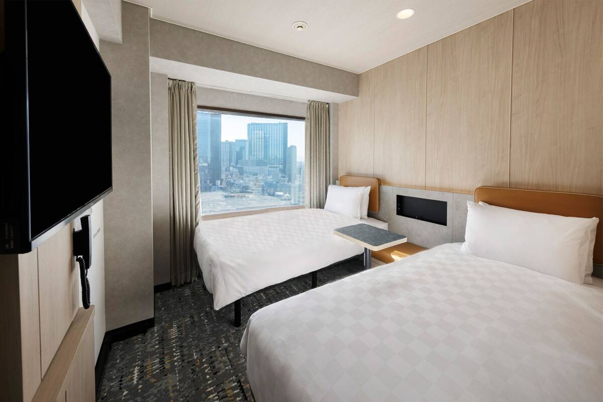 A hotel room with two beds, a TV, a small table, and a view of the city.