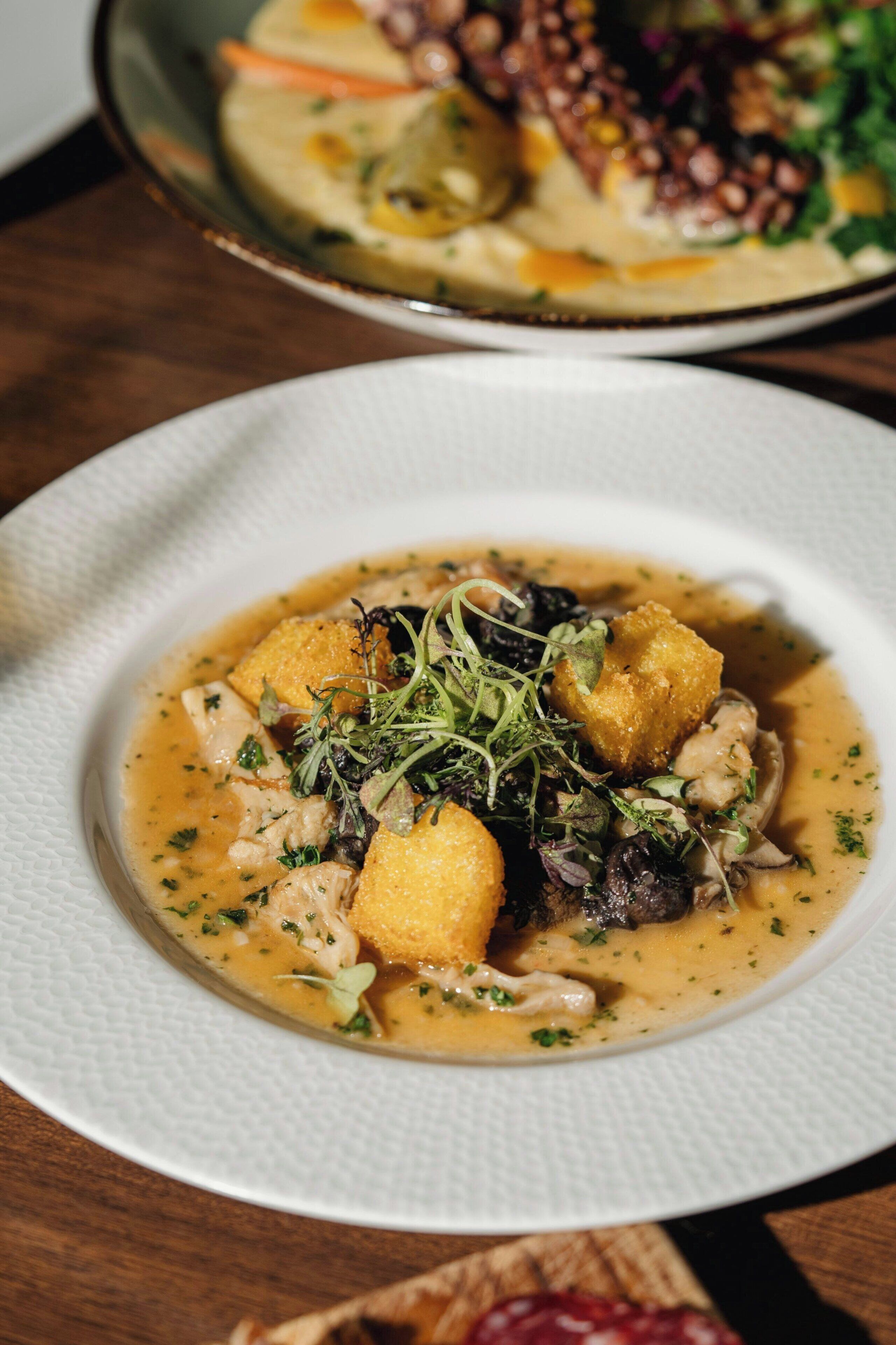 Un piatto bianco con una zuppa cremosa, guarnita con verdure e un elemento dorato-brunastro, su un tavolo di legno.