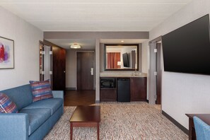 A hotel room with a sofa, a coffee table, a flat-screen TV, a kitchenette, and a door leading to another room.
