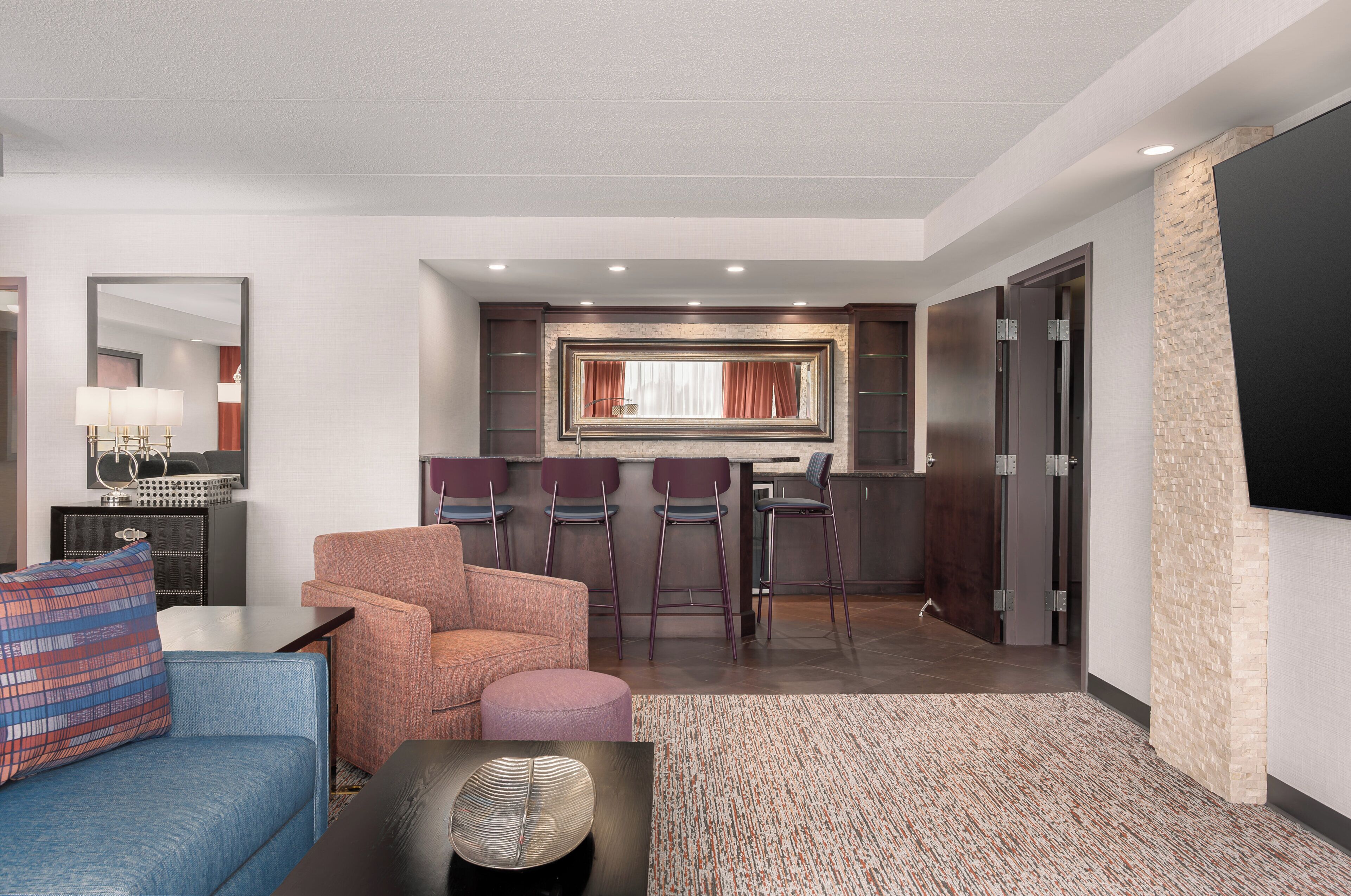 A hotel room with a seating area, a kitchenette, and a large mirror.
